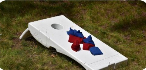 White cornhole board on a grassy lawn with red and blue bean bags resting on its surface.
