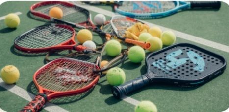 Tennis court covered with scattered rackets, pickleball paddles, and lots of yellow tennis balls.