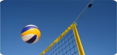 Volleyball in mid-air near a yellow beach volleyball net against a clear blue sky.